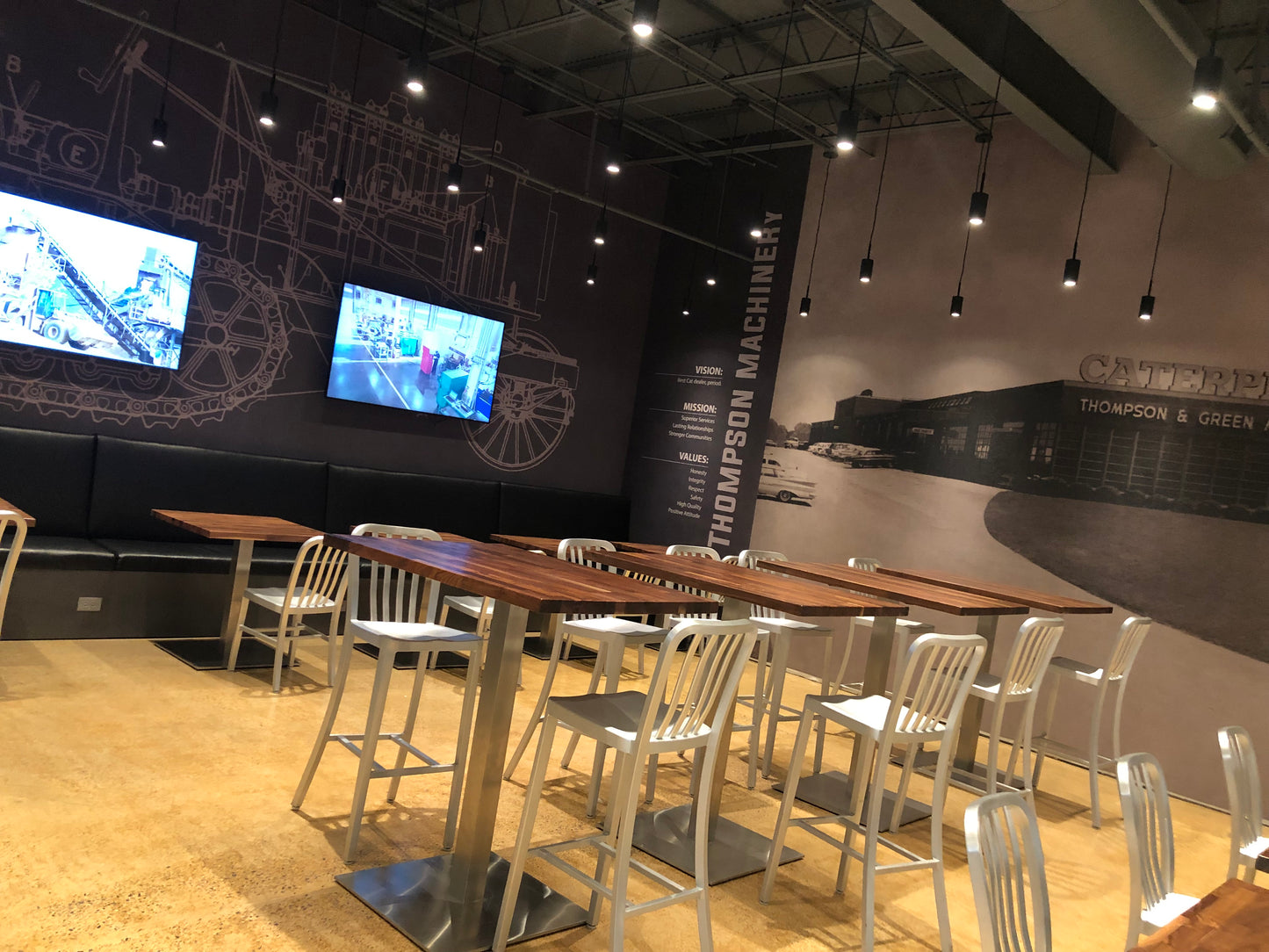 Modern dining area with wooden tables and metal chairs against a wall with large screens and branding.