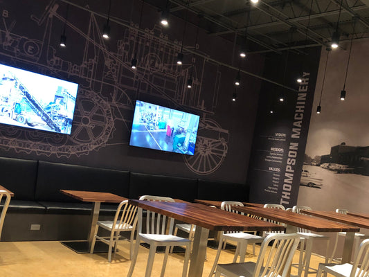 Modern dining area with wooden tables and metal chairs against a wall with large screens and branding.